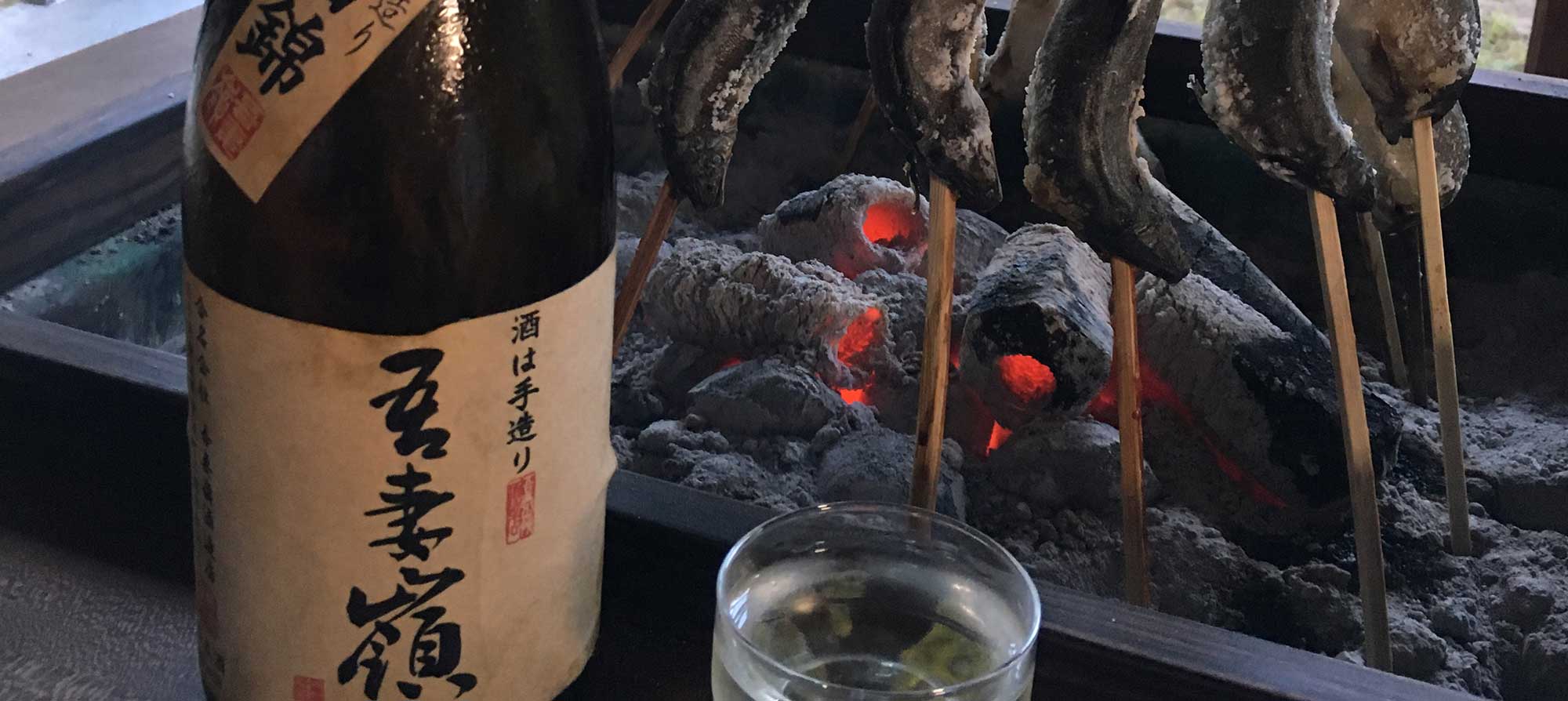 Рыба жарится на палочках в традиционном японском очаге. В стопку налито сакэ